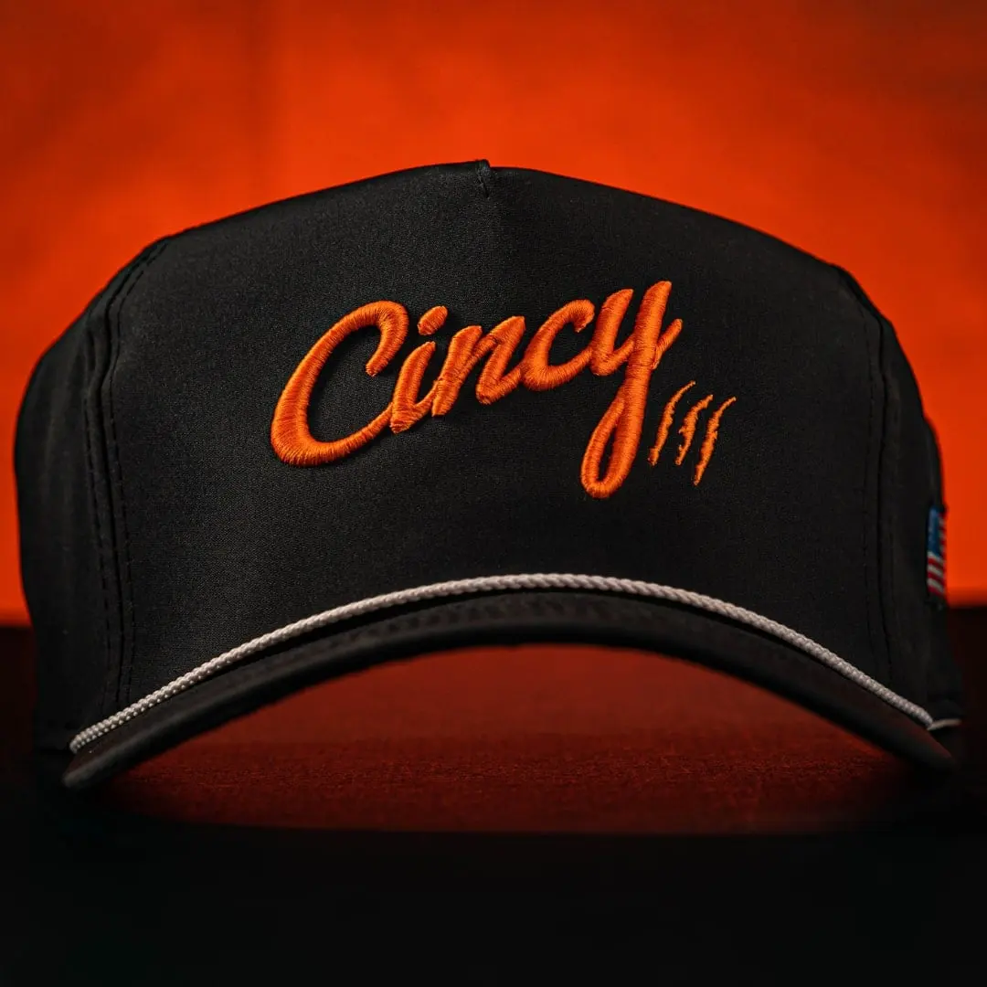 Black Cincy hat with orange script and a white rope, lit against a glowing orange backdrop