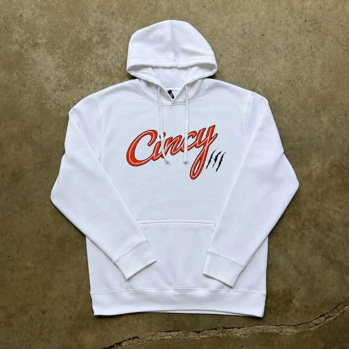 White hoodie with orange Cincy script outlined in black and claw slashes, laid flat on a concrete floor