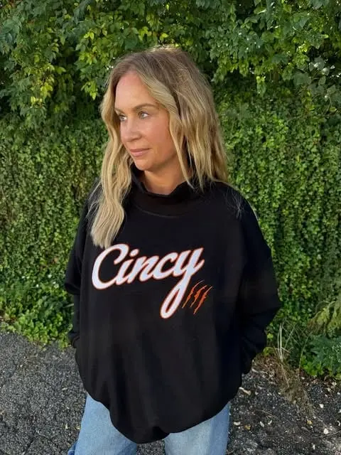 A woman in a black Cincy crewneck sweatshirt standing in front of a green hedge