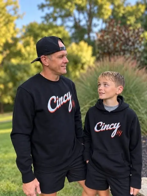 A man in a black Cincy crewneck and a boy in a black Cincy hoodie smile at each other outdoors in evening light