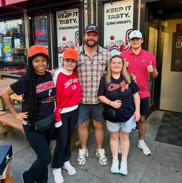 Five friends in Cincy hats and Cincinnati gear posing outside a restaurant, two of them in orange Cincy hats