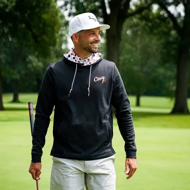 A smiling golfer in a black Cincy hoodie with a patterned hood lining and a white cap, walking a fairway with a club