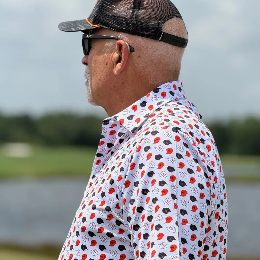 A man in sunglasses and a mesh cap wearing The Cincy Hat Pattern Polo, printed with small red, black and white hats, beside a golf course pond