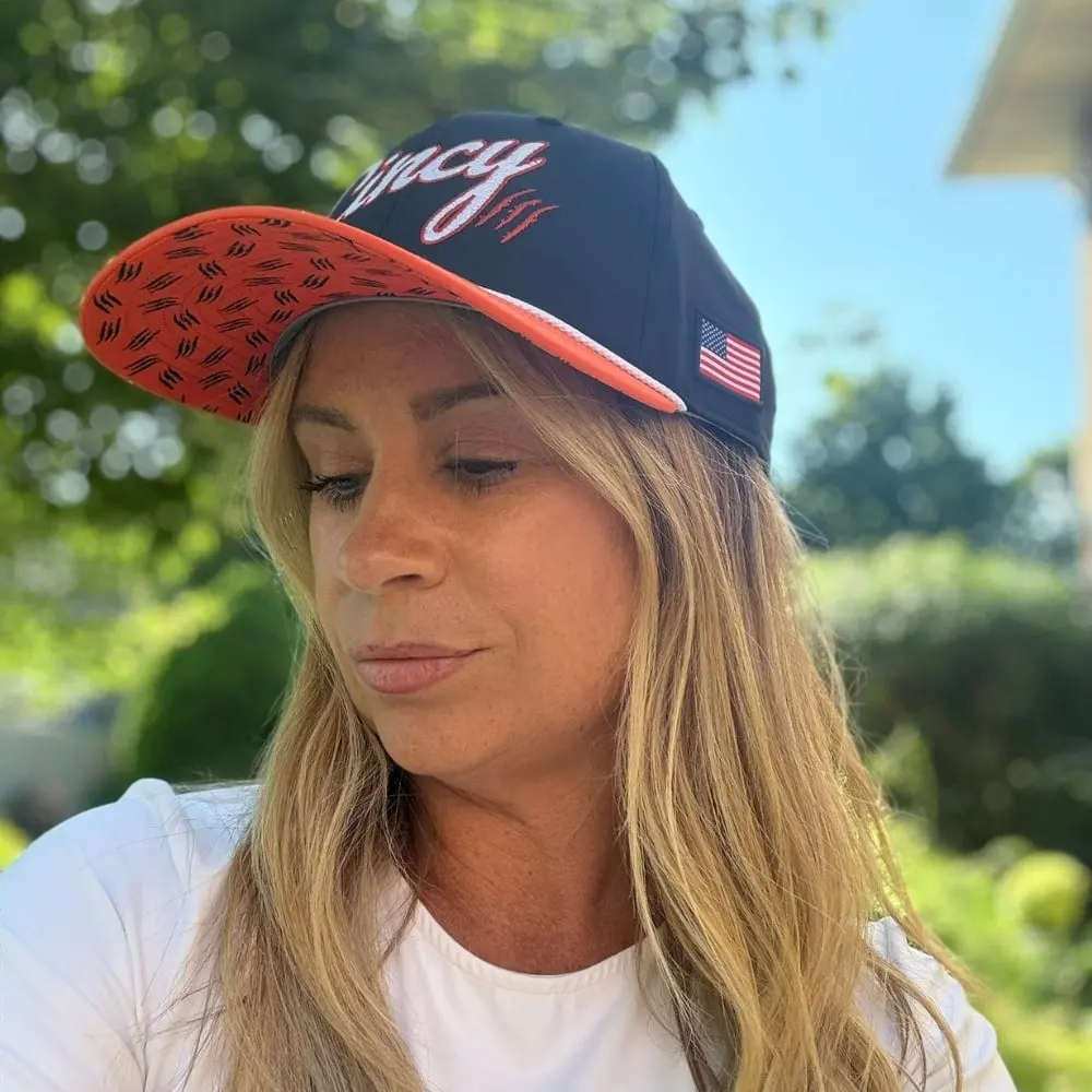 A woman outdoors wearing The Training Camp Mid-Crown, a navy hat with white Cincy script, a patterned orange brim and a flag patch