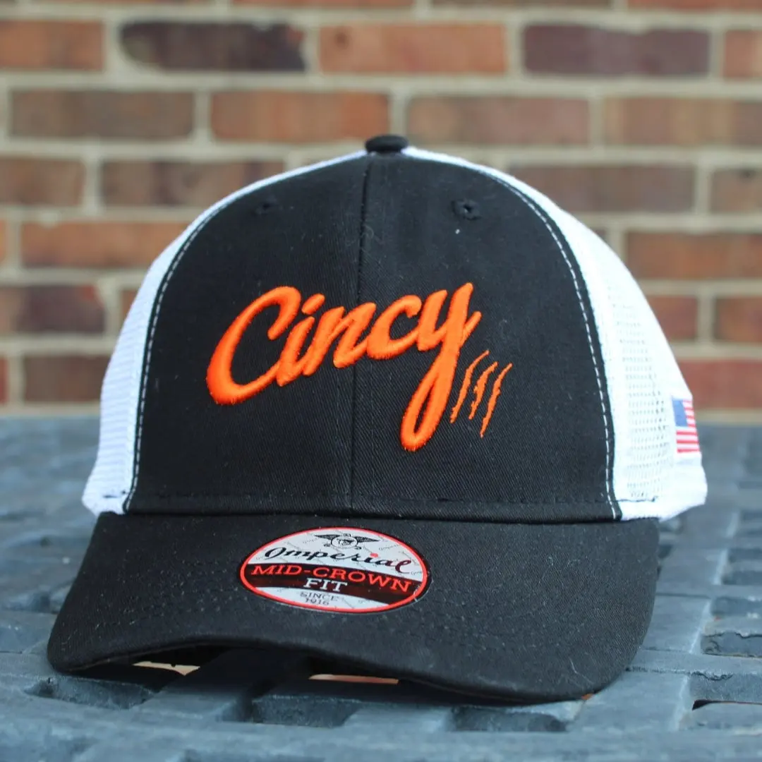 Black mesh-back hat with orange Cincy script and a white mesh back, resting on a ledge in front of a red brick wall