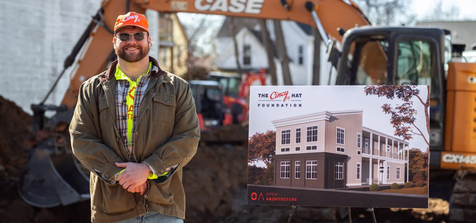 Ted Karras in an orange Cincy hat and a hi-vis vest at the Madisonville groundbreaking, beside a sign showing a rendering of the new building, with an excavator behind him